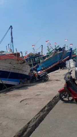 The video shows a harbor scene with several boats docked. In the foreground, there are two motorcycles parked on a concrete surface. The boats are varied in color and condition, with some appearing well-maintained and others showing signs of wear and tear. One boat has a colorful hull with red, yellow, green, and blue stripes. Another boat, docked next to it, is partially obscured and appears to be in disrepair with peeling paint. Indonesian flags are visible on some of the boats. The sky is clear and blue, suggesting a sunny day. The overall impression is of a working harbor with fishing vessels.