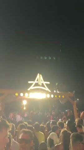 This is a vertical video of a nighttime outdoor concert or party. A crowd of people is gathered in front of a stage or performance area. Above the stage is a large, illuminated sign with the letters "AA" stylized within a triangle. The stage is lit with bright lights, and there appears to be a DJ booth or performance setup visible. Smoke or fog is present in the air, illuminated by the stage lights. The crowd is diverse, with many people holding up their phones to record or take pictures. The overall atmosphere appears to be energetic and festive.