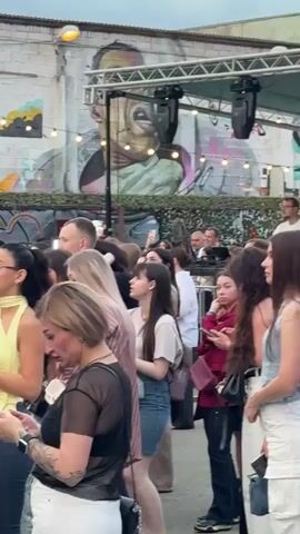 A crowd of people are gathered outdoors, seemingly at an event or concert. The background features a building with graffiti art on the wall and string lights overhead. Some individuals are holding phones, suggesting they are recording or taking pictures. The focus shifts to a stage area where a DJ is performing, with a large sound system and lighting equipment visible. The DJ, wearing a white shirt and sunglasses, is interacting with the audience. The atmosphere appears lively and energetic.