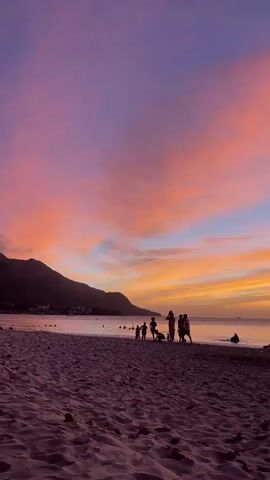 A group of people are gathered on a beach during sunset. The sky is filled with vibrant colors of pink, orange, and purple. The sand on the beach is a deep purple hue. In the distance, a mountain range is visible on the left side of the frame. Several individuals and a child are silhouetted against the colorful sky, some appearing to be playing or walking on the beach. A few people are in the water, and one person is seated on the sand in the foreground, facing away from the camera. The ocean reflects the colors of the sunset.