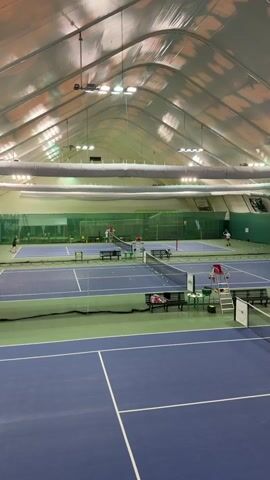 The video shows an indoor tennis court with a high, arched ceiling. Two blue courts are visible, with green surroundings. There is a net separating the two courts. Balls are scattered across the far court. Two people are playing tennis on the far court. The camera is stationary and is positioned to give a wide view of the courts. The lighting in the room is bright. A small white table, a chair, and a ladder are positioned next to the near court. Benches are on each side of the nets.