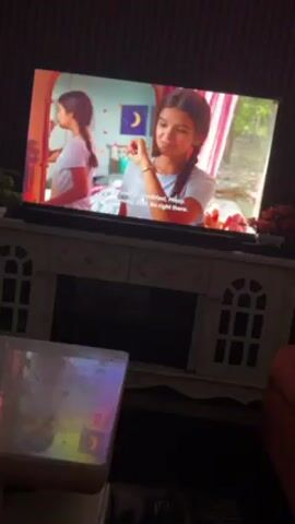 Here is a description of the video:

The video shows a TV screen displaying a scene from a TV show or movie. The scene alternates between a young woman in a bedroom and a man in a kitchen, both characters appearing to be preparing for the day. The TV is sitting on top of a white TV stand or console table, and there is a glass-topped coffee table in front of it. The room is dimly lit. The TV show has subtitles. The setting appears to be a domestic living room.