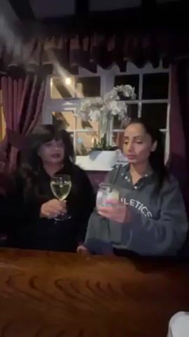 Two women are sitting at a wooden table inside a dimly lit establishment, possibly a restaurant or a pub. The woman on the left has dark hair and is wearing a dark colored outfit. She is holding a wine glass filled with a light-colored liquid, presumably white wine. The woman on the right has her dark hair pulled back into a ponytail and is wearing a grey sweatshirt with the word "Athletics" visible. She is holding a glass with a light-colored mixed drink with a straw. Both women are facing each other and appear to be engaged in a conversation. In the background, there is a window with white framing and a flower arrangement on the windowsill. The room is decorated with dark curtains and wood paneling, contributing to the intimate atmosphere.