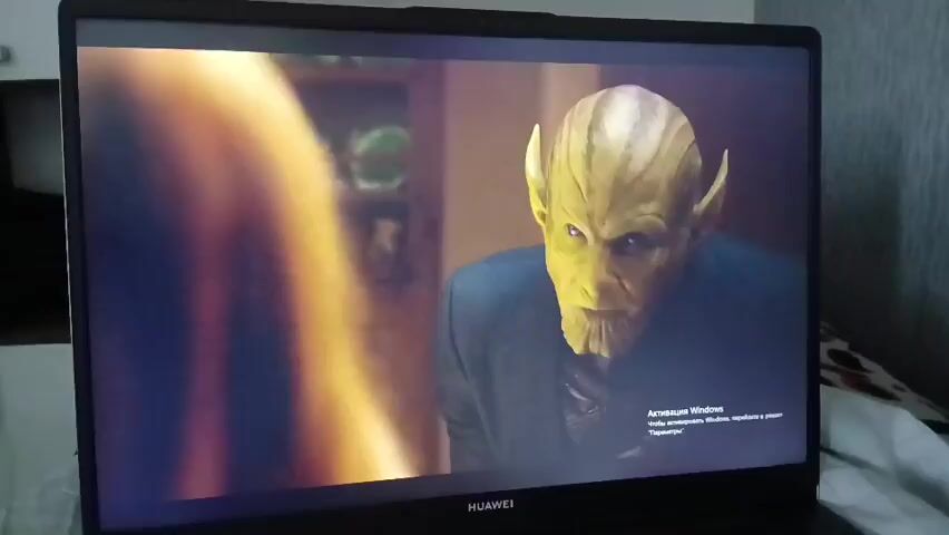 THE VIDEO DISPLAYS A SCENE FROM A MOVIE OR TELEVISION SHOW PLAYING ON A LAPTOP SCREEN. ON THE LEFT SIDE OF THE SCREEN, A WOMAN WITH SHORT, BLONDE HAIR WEARING A WHITE T-SHIRT WITH A BLACK LOGO THAT APPEARS TO READ "NIN" IS VISIBLE. SHE HAS A SERIOUS EXPRESSION. ON THE RIGHT SIDE OF THE SCREEN, A MALE CHARACTER WITH YELLOWISH-GREEN SKIN AND DISTINCT EARS, WEARING A DARK JACKET, IS SHOWN. THE BACKGROUND APPEARS TO BE AN INDOOR SETTING WITH A DOOR AND FURNITURE VISIBLE. IN THE BOTTOM RIGHT CORNER OF THE LAPTOP SCREEN, A WINDOWS ACTIVATION WATERMARK IS PRESENT. THE LIGHTING IS DIM, WITH SOME GLARE FROM THE LAPTOP SCREEN. THE VIDEO APPEARS TO BE A SCREEN RECORDING OR A PHOTO TAKEN OF THE LAPTOP SCREEN.