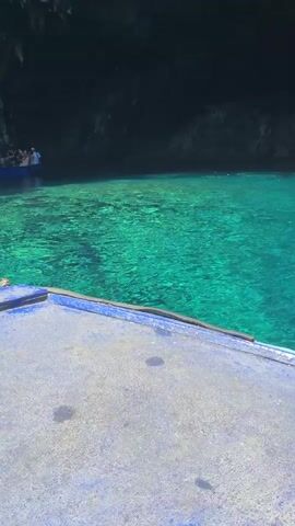 A boat with blue trim approaches a cave opening with turquoise water. The boat is moving from right to left. Inside the cave, more boats with people can be seen. The water is clear and the cave walls are rocky and dark. Sunlight filters into the cave, illuminating parts of the water and the rock formations. The scene transitions to an underwater view where fish are visible swimming in the bright turquoise water. The cave walls are also visible from below the water's surface, with sunlight beaming down. The video ends showing the cave walls and a glimpse of trees at the top.