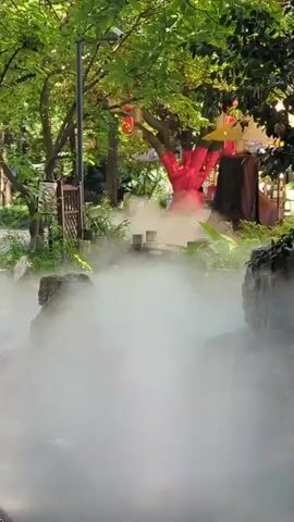 The video shows a park-like setting with lush green trees and foliage. A pathway is visible, with decorative wooden fences and rocks along its sides. A significant amount of mist is present, creating a diffused and ethereal atmosphere. In the background, a tree with red illumination stands out, and red lanterns are hanging from branches. Several structures, possibly stalls or gazebos, are also visible. A few individuals are present in the background, one walking near a stall and another standing. The lighting suggests it might be late afternoon or early evening.