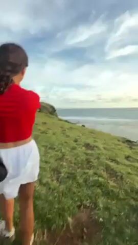 The video shows a woman walking on a grassy hill overlooking a beach at sunset. The woman is wearing a white dress and white sandals. She is carrying a white bag over her shoulder and holding something in her hand. The sun is setting over the ocean, casting a golden glow on the scene. There are other people visible on the hill and on the beach below. The sky is partly cloudy. The camera pans to show the entire hillside and coastline.