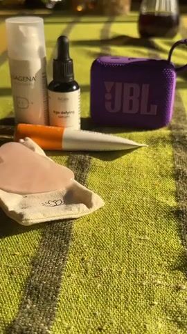 The video shows a static close-up of several items on a green and yellow striped tablecloth, including a bottle of "COLLAGENA PROFESSIONAL IDENTITY" cream, a bottle of "BioDeana Age defying serum," an orange tube of cosmetic product, a white tube of cosmetic product, a heart-shaped rose quartz Gua Sha tool in a small white bag, and a purple JBL speaker. The scene then cuts to a close-up of a folded white napkin on a white plate, followed by a shot of a brown dog asleep on a red rug. Part of a chair leg and a yellow dog collar are also visible in this last frame.