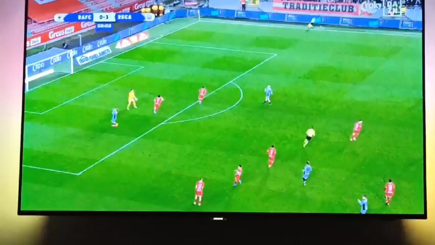 The video shows a soccer game on a Philips television screen. The score is 0-3, and the game is in the 58th minute. The screen shows a player in a light blue jersey celebrating, followed by an overhead shot of the game in progress. There are several players in light blue and red jerseys on a green field. There is a referee in black. The stadium is visible in the background. There are advertisements visible around the field. The ambient lighting around the television is a rainbow of colors.