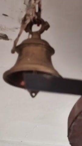 A brass bell hangs from a rope attached to the ceiling. A larger, ornate bell is visible to the right. The bells are likely in a temple or religious setting.