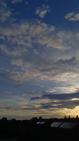 The video shows a static view of a sunset over a silhouetted cityscape. The sky is a gradient of blue, with scattered white and gray clouds. The sun is a bright orange glow on the horizon, partially obscured by darker clouds. Below the sky, the silhouettes of buildings, rooftops, and trees are visible. Some of the rooftops appear to have solar panels. The overall scene is dark and atmospheric, capturing the transitional light of dusk.