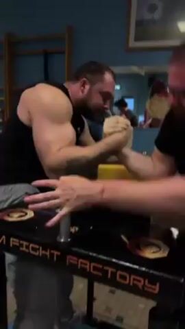 Two men are arm wrestling in what appears to be a gym. One man is wearing a black tank top and gray sweatpants, while the other is wearing a black t-shirt and glasses. They are at an arm wrestling table with the words "FIGHT FACTORY" written on it. The man in the black tank top appears to be winning, as he pushes the other man's hand down. There are other people in the background, and gym equipment such as wooden ladders along the wall.