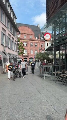 A man walks down a cobblestone street, pulling a black electric scooter behind him. He is wearing a grey t-shirt and black pants. He passes a woman in a grey t-shirt and black pants, who is walking her dog. In the background, several other people are walking on the street. The street is lined with buildings on both sides, and there are shops and restaurants. The sky is blue with some clouds.
