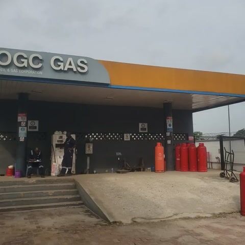 The video shows a gas station with several red propane tanks stacked in the foreground. A tall telecommunications tower is visible in the background, along with a white building and some trees. The sky is overcast, and the ground is paved with concrete. In the first clip, a sign that says "GAS" is visible on the side of a building. In the second clip, more propane tanks are visible, along with a chain-link fence and a metal pole.