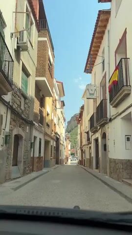 The video shows a narrow street in a town. The street is lined with buildings on both sides. The buildings are two or three stories tall and appear to be made of brick and plaster. The street is paved with asphalt. In the distance, a mountain can be seen. The sky is clear and blue. The video is shot from a car that is driving down the street. The car appears to be moving at a slow speed. There are no people or other vehicles visible in the video.