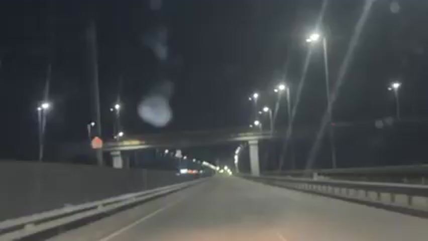 The video shows the perspective of someone driving on a bridge at night. Streetlights line both sides of the road, and an overpass is visible above. The road is empty of other vehicles. The image is somewhat blurry.