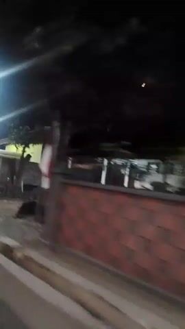 The video shows a nighttime scene from a moving vehicle. The camera pans across a dimly lit area with some structures and a bright overhead light. There is a red bin visible in the foreground. The vehicle then passes by a building with a green wall and a large dark shutter or door. Further on, a house with its lights on is visible, with a vehicle parked in front of it. The overall environment appears to be a street or road in a residential or semi-commercial area at night.