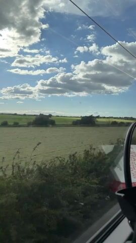 The video shows a view from inside a vehicle, likely a train or a car, looking out at a landscape. The sky is a vibrant blue with scattered white cumulus clouds, some of which are partially lit by sunlight. In the distance, there are rolling green fields and trees. A portion of the vehicle's interior is visible, including a black object that may be a rearview mirror or a seat. The overall scene suggests a rural setting on a bright, cloudy day.