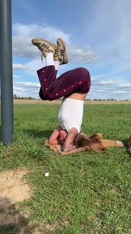 A woman is performing a headstand on a blanket in a grassy field. She is wearing burgundy pants with white stripes, a white t-shirt, white socks, and brown sneakers. She has long brown hair and visible tattoos on her arms. Behind her are several gray metal poles forming a pull-up bar structure. The ground around her is a mix of grass and dry, cracked earth. In the background, there is a field with scattered trees under a blue sky with scattered white clouds. The camera pans from the woman doing the headstand to a wider view of the pull-up bars and the field.