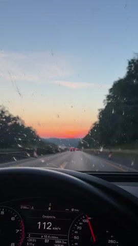The video begins with a view from inside a car, showing the dashboard and the road ahead. The sun is setting, casting a warm orange and pink glow across the sky. The car is moving at a speed of 111 km/h, indicated on the dashboard. The road is a highway with multiple lanes, and there are other cars in front. Trees line the sides of the road, and hills are visible in the distance. The windshield is slightly dirty, with some streaks and smudges. The overall impression is that of a peaceful drive on a highway at sunset.