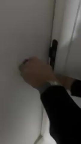 A person wearing a black long sleeve shirt and a watch opens a white door. A small, beige, fluffy dog jumps up towards the person, who steps into the room, wearing brown sandals and blue jeans. The room has grey tiles and a welcome mat. The dog continues to jump and moves around excitedly near the person's feet.