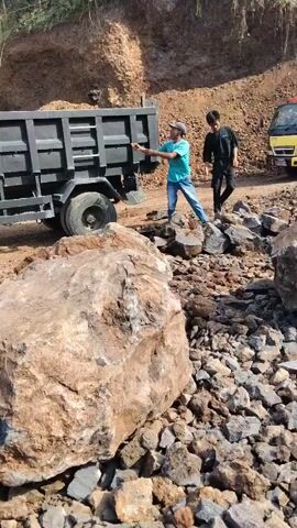 Two individuals are working near a large dump truck and piles of rocks. The person in the blue shirt and hat appears to be operating a jackhammer on a rock, while the person in the black shirt is observing and may be assisting with the rocks. The environment is an outdoor site with a dirt hill in the background. The scene suggests construction or demolition work involving the breaking and loading of large rocks.