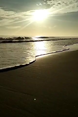 The video shows a beach with small waves coming in. The sun is shining brightly in the sky, reflecting off the water. The sand appears to be a dark color. There are some clouds in the sky.