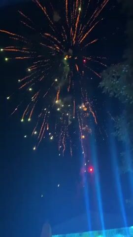 This is a vertical video taken at night during a fireworks display. The sky is dark with some smoke from the fireworks. Several bursts of fireworks are visible, with bright yellow and red sparks radiating outwards. In the lower portion of the screen, there are bright blue spotlights directed upwards and to the right. A silhouette of a tree or leaves is visible on the right edge of the frame. The bottom of the screen shows a blurred view of what appears to be a crowd of people gathered, with some blue lights illuminating them. The video captures the dynamic and festive atmosphere of a fireworks show.