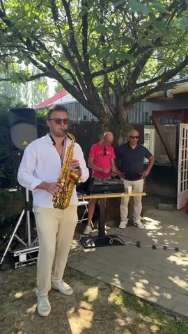 The video opens with a close-up of a man playing a saxophone in an outdoor setting. The camera then pans to the left, showing a hedge and a speaker. The scene shifts to show people sitting on chairs outdoors, with a woman in a pink dress in the foreground. The camera moves to show other guests, including a woman adjusting her clothing and a man with glasses. Next, the video shows a group of men sitting on chairs and two women standing and talking. The scene changes again to show a table set for a party with a candelabra and various items on it, with people standing in the background. The video appears to capture a social gathering outdoors.