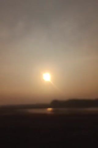 A video shows a sunset over a body of water. The sun is visible in the sky, and its rays are emanating outwards. The water appears dark, and there is a silhouette of trees on the horizon. The sky is hazy, with a gradient of colors from orange to gray.