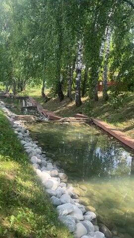 A stream flows through a wooded area. The stream has a rocky bed and is lined with stones. Wooden planks are placed across the stream at intervals, creating small dams or water features. Tall trees with green foliage line the banks of the stream, casting shadows on the water. The scene is well-lit by natural sunlight. There are no people or animals visible in the video. The overall impression is one of a natural, peaceful environment, possibly a park or a landscaped garden.