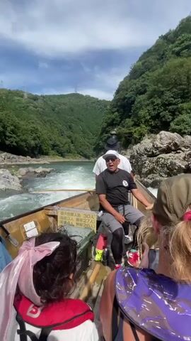 A group of people are on a wooden boat traveling down a river. The boat is moving fast, creating white water. The river is surrounded by lush green mountains. There are two men at the front of the boat, one steering and one standing behind him. The steering man is wearing a black t-shirt, grey shorts, and a white hat. He is smiling and appears to be talking to the passengers. The other man is wearing a white t-shirt and a black hat. Several passengers are visible in the boat, including two women with their backs to the camera, one wearing a pink bow in her hair and the other a tan hat. The woman with the tan hat is holding up a phone to take a picture. There are also signs in Japanese on the side of the boat. The sky is blue with some clouds.