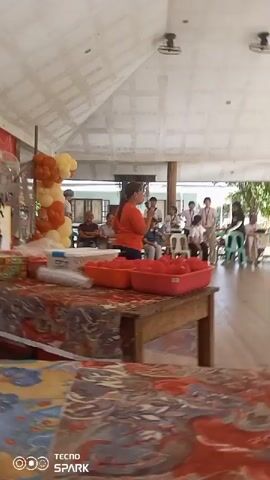 A woman in an orange shirt stands facing a seated audience, holding a microphone and speaking. In front of her is a table covered with a patterned cloth, displaying several items including plastic containers filled with red items, possibly fruit, and a roll of clear plastic wrap. To her left, a balloon arch with orange and yellow balloons is visible. The scene appears to be an outdoor or semi-outdoor event, with a covered ceiling supported by wooden beams and the presence of natural light. In the background, several other people are seated and standing, some of whom appear to be younger individuals. The overall environment suggests a community gathering or a presentation.