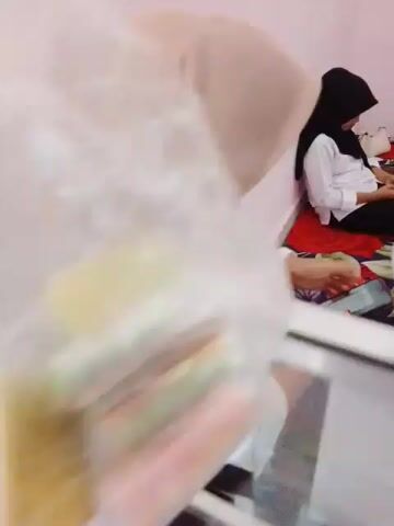 The video opens with a close-up of a clear plastic bag filled with colorful, rolled-up candies, possibly ice pops or a similar frozen confection. The camera then pans to reveal a room with light pink walls. In the background, two young women are seated on the floor, which is covered by a colorful mat with floral and leaf patterns. One woman, on the left, is wearing a black hijab and a white shirt, and is looking at her phone. The other woman, on the right, is wearing a light brown hijab and a white shirt, and is smiling and looking to her right. The camera then moves to focus on the woman in the light brown hijab, who is wearing glasses and a white shirt. She appears to be holding a phone and looking down at it. A ceiling fan is visible on the wall above and behind the seated women. Further to the right, there is a red shelf unit with various items on it, including what appears to be a laptop and bottles. Next to the shelf unit is a glass display case filled with items, possibly clothing or accessories, and some items are on top of the case. The video ends with a shot of the woman in the light brown hijab, her face partially obscured by her hijab.