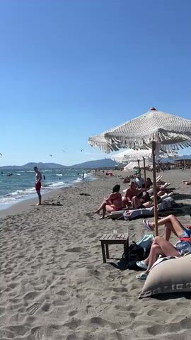 The video shows a sandy beach with the ocean on the left and people relaxing on the right. A man in red shorts stands near the water's edge, looking out at the sea. In the water, several people are visible, and a large number of kites are flying in the air above the ocean, suggesting a windy day suitable for kite surfing. On the right side of the beach, people are seated on lounge chairs under a large, white umbrella with fringe detailing. A small wooden table is positioned between two lounge chairs. The sky is clear and blue, with the sun shining brightly. The overall scene depicts a lively beach environment on a sunny day.
