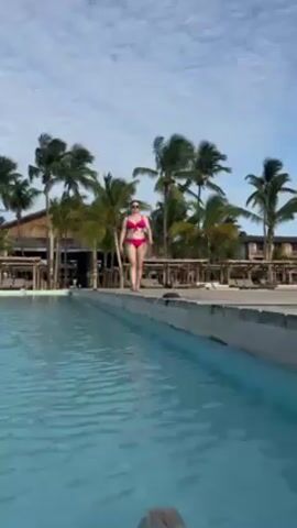 A woman wearing a pink bikini walks along the edge of a swimming pool. The pool has light blue water, and there are palm trees and a wooden structure in the background. The woman is walking towards the camera, and the sky above is blue with white clouds.