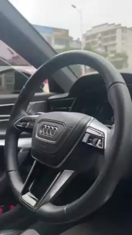 The video shows the interior of a modern car, likely an Audi, from the driver's perspective. The camera pans from the steering wheel to the dashboard and center console. The steering wheel is black with the Audi logo prominently displayed in the center. The dashboard features a digital display, and the center console has a large touchscreen. A small decorative item hangs from the rearview mirror. The person in the driver's seat is wearing khaki pants. Through the car window, buildings and traffic are visible. The weather appears overcast.