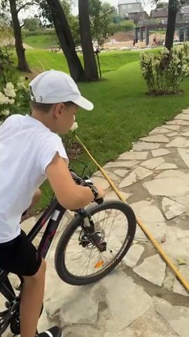 A boy in a white t-shirt and cap rides a black bicycle on a paved path in a park. He is facing away from the camera. Other people are walking in the background, and there is a grassy area with trees. The path is made of irregularly shaped stones.