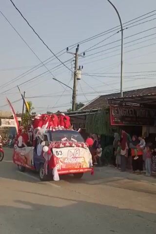 A group of people, mostly women wearing red outfits and headscarves, are riding on and in a blue and white decorated pickup truck. The truck is adorned with red and white streamers, balloons, and banners. Some individuals are holding pom-poms and small Indonesian flags. The truck has "LA AEROBIC" visible on its front. A banner on the side of the truck has "INDONESIA ADIL & MAKMUR" written on it. The truck is moving down a street lined with buildings, including one with a sign that reads "DENGKEL MOTOR IK" in the background. Several onlookers are standing on the side of the street, watching the procession. Two motorcycles are visible to the left and right of the truck. The sky is clear and the sun is shining.