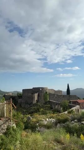 The video shows a pan across a landscape. The video starts by showing a ruined stone structure, possibly a castle, on a hill. The camera pans to the right to show a town with houses and trees, with mountains in the background. The sky is partly cloudy, and the sun shines brightly. There is the sound of a dog barking in the background.