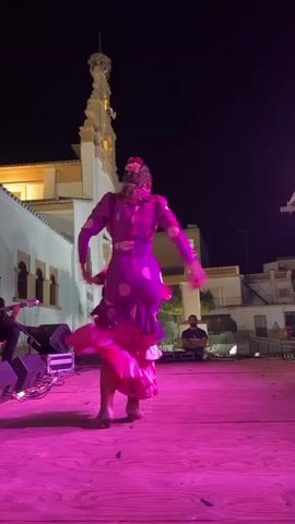 A woman in a vibrant purple polka-dotted dress with ruffled pink accents performs a flamenco dance on a wooden stage under a night sky. The stage is illuminated by bright pink lights. In the background, a building with architectural details and a steeple is visible, along with other structures and a few people observing the performance. To the left of the stage, audio equipment, including speakers, is set up. A person is playing a guitar to the left of the frame. The dancer is captured mid-motion, her dress swirling around her as she moves.