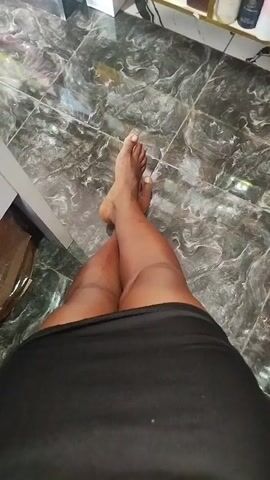 The video shows a close-up, top-down view of a person's legs and feet. The person is wearing a black skirt that covers their thighs. Their legs are crossed at the knees, and their bare feet are visible. The floor appears to be tiled with a dark, marbled pattern. In the background, out of focus, are shelves with various items that appear to be bottles or containers. The lighting is somewhat dim. The video progresses with slight camera movements, shifting focus and angle on the legs and feet.