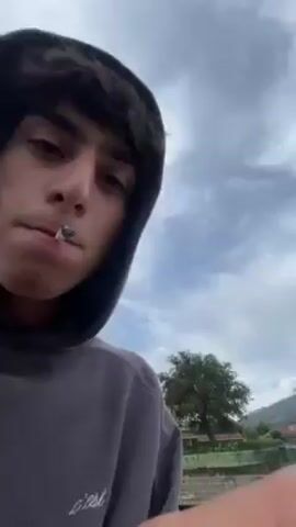 The video opens with a close-up of a young man with dark hair and a gray hoodie, smoking a cigarette. The background shows a cloudy sky and some trees. The camera then shifts to another young man, sitting on a ledge overlooking a body of water. He is wearing a white sweatshirt with a red anchor symbol and purple pants. The camera follows a thrown object, appearing to be a rock, which splashes into the water.