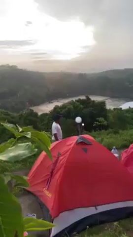 The video shows a campsite overlooking a coastal scene. There are three tents, two red and one blue-green, set up on grassy ground. A man stands near one of the red tents, looking out at the view. The landscape includes a beach, ocean with waves, rocky cliffs, and greenery. The sky is overcast, with a hint of sunlight breaking through the clouds. There is a small light fixture nearby. A water bottle sits by one of the red tents. The tents appear to be located on a slight incline overlooking the beach and ocean.