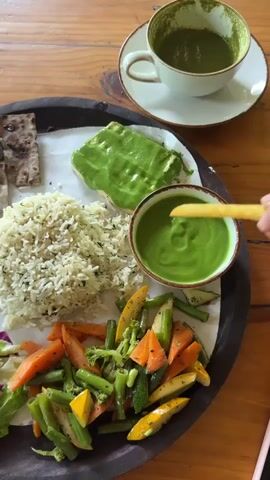 A high-angle shot shows a wooden table with a large, round, wooden platter. On the platter, there is a variety of food items. There's a portion of cooked rice mixed with herbs, a few pieces of thin, grilled bread, a slice of something covered in a vibrant green sauce, a small bowl filled with the same green sauce, and a colorful mix of sauteed vegetables. A small salad with lettuce and red cabbage is also visible. A hand is dipping a fry into the bowl of green sauce. To the side of the platter, there's a white teacup with a saucer, containing a green liquid. The table surface is wooden. The lighting suggests an indoor setting.