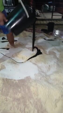 A person wearing blue and green patterned shorts is barefoot and pouring a dark liquid from a black bucket onto a large mound of light-colored powders. The person steps into the powder, sinking their feet into it. The liquid creates dark lines and puddles on the surface of the powder. A black cable or rope lies across the powder. In the background, a pickup truck, a tile floor, and other individuals are visible.