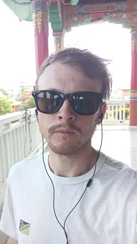 A man with short brown hair, a mustache and a sparse beard stands in front of a wall with colorful murals. He wears a plain white shirt, black sunglasses and black earphones with a cord. He looks directly at the camera and speaks. The camera is positioned at a close-up angle, capturing his face and upper torso. The background includes a view of the exterior of a temple-like structure, adorned with elaborate paintings and architectural details. The colorful murals feature various figures and symbols, likely religious in nature. The man appears to be recording a video or giving a presentation, possibly sharing his experiences at the location.