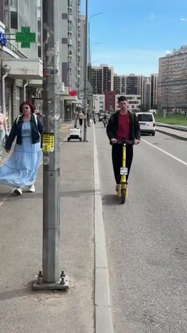 The video captures a bustling street scene in Murino, Russia, on a bright, sunny day. A person rides a yellow electric scooter down a paved road, passing several other individuals and vehicles. In the foreground, a woman in a light blue dress walks along the sidewalk next to a grey pole adorned with stickers. Behind her, shops line the street, with more people walking by and a white cart on wheels. Further down the road, several motorcycles and a white van can be seen. Tall apartment buildings form the backdrop of the urban environment. The overall atmosphere appears to be lively and typical of a summer day in a residential city.

Visible text includes advertisements on stickers on the pole, and license plates on the scooter and vehicles, though the text is not clearly legible. The green cross symbol on a building in the background might indicate a pharmacy or medical facility. The sun is high in the sky, suggesting it is likely daytime.