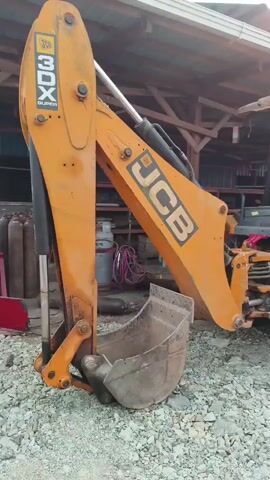 A yellow JCB backhoe loader is parked outdoors on a gravel surface under a wooden structure with a corrugated metal roof. The front bucket of the loader is visible. The video pans from the front of the loader to the rear, showing the cab and the large rear tires. In the background, a person walks away from the camera along a path next to a row of buildings. A gas cylinder and hoses are visible on the left side of the frame.