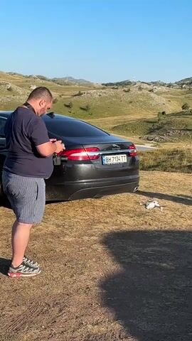 A man is standing outdoors next to a black car, operating a drone. The drone is on the ground a few feet away from the car. The man is wearing a dark blue t-shirt, striped shorts, and sneakers. The car has a license plate that reads "BH 7154 TT". The background is a hilly, dry landscape under a clear blue sky. The sun is shining.